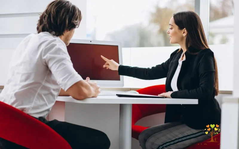 A man and woman seated at a table, engaged in discussion while looking at a computer screen.